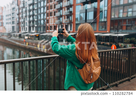 Back view of young European tourist makes photo or video on smartphone at city of Gdansk, Poland. Hipster girl with backpack hold on smart phone gadget outdoors Back view of young European tourist makes photo or video on smartphone at city of Gdansk, Poland. Hipster girl with backpack hold on smart phone gadget outdoors 110282784