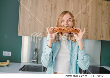 Portrait of a hungry young woman eating wholesome sandwich, having tasty breakfast in a kitchen at home in the morning Portrait of a hungry young woman eating wholesome sandwich, having tasty breakfast in a kitchen at home in the morning 110284208