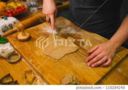 Female hand cut gingerbread dough with a sharp knife at kitchen. Traditional Christmas home bakery. Baking, cooking and christmas concept Female hand cut gingerbread dough with a sharp knife at kitchen. Traditional Christmas home bakery. Baking, cooking and christmas concept 110284240