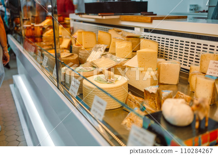 Delicatessen shop with various cheese in the display cabinet and a worker behind the counter at city market Delicatessen shop with various cheese in the display cabinet and a worker behind the counter at city market 110284267