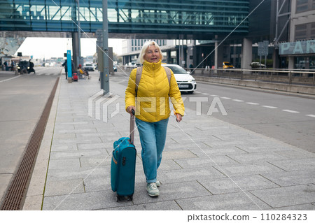 Happy attractive 60s senior female traveler walking with a suitcase at the modern transport stop outdoors near the modern airport terminal Happy attractive 60s senior female traveler walking with a suitcase at the modern transport stop outdoors near the modern airport terminal 110284323