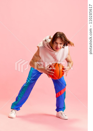 Young man basketball player in retro sport uniform stands on attack training technique holding ball against pastel pink studio background. Young man basketball player in retro sport uniform stands on attack training technique holding ball against pastel pink studio background. 110285617
