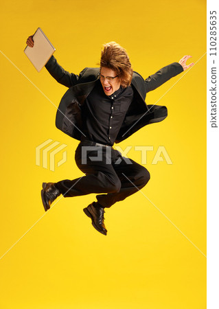 Young man, dressed smart casual, fashion and glasses outfit jumping of joy and raising hands holding laptop against yellow background. Young man, dressed smart casual, fashion and glasses outfit jumping of joy and raising hands holding laptop against yellow background. 110285635