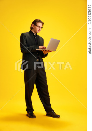 Full length portrait of young confident man, student holding laptop and having video call against vivid yellow background. 110285652