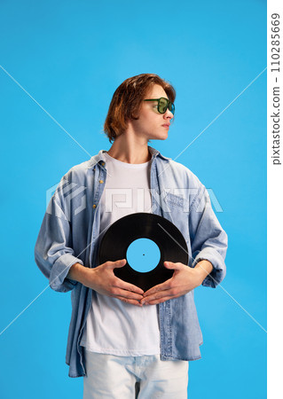 Guardian of music. Portrait of cool attitude man carefully holding vintage vinyl record and looking away against blue background. Guardian of music. Portrait of cool attitude man carefully holding vintage vinyl record and looking away against blue background. 110285669