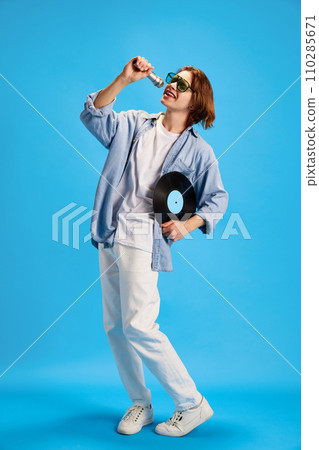 Young man wearing fashion casual outfit and sunglasses singing retro microphone and holding vintage vinyl record disk against blue studio background. Young man wearing fashion casual outfit and sunglasses singing retro microphone and holding vintage vinyl record disk against blue studio background. 110285671