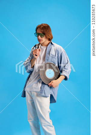 Young man wearing fashion casual outfit and sunglasses, looks like pop star, singing retro microphone and holding vintage vinyl record disk. Young man wearing fashion casual outfit and sunglasses, looks like pop star, singing retro microphone and holding vintage vinyl record disk. 110285673