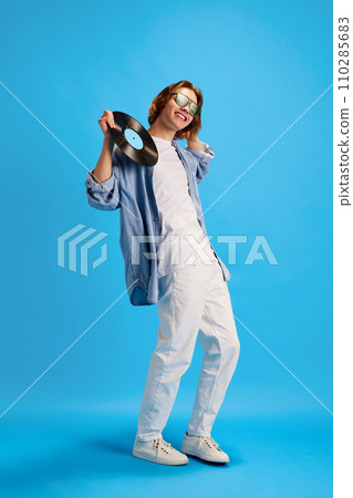 Full length portrait of young man, hipster in stylish sunglasses posing with vinyl record, disk against blue studio background. Concept of hobby. 110285683