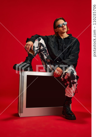 Young man in style clothes, looks as cowboy sitting on retro TV and looking away against red studio background. Concept of people and technology. Young man in style clothes, looks as cowboy sitting on retro TV and looking away against red studio background. Concept of people and technology. 110285706