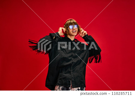 Portrait of young guy, dressed retro outfit, looks like cowboy enjoying listening music in headphones against vibrant red background. Concept of self-expression. 110285711