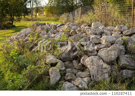 Large stones lie in a large pile on the grass. Beautiful large stones stacked in the grass. Rock stones are dumped into a large pile in the green grass. 110287411