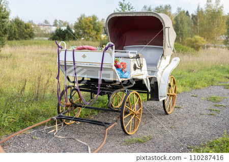 A beautiful empty carriage stands without a horse on a rural road. The elegant white carriage stands empty. Concept of an old fashioned carriage driven by a horse. A beautiful empty carriage stands without a horse on a rural road. The elegant white carriage stands empty. Concept of an old fashioned carriage driven by a horse. 110287416