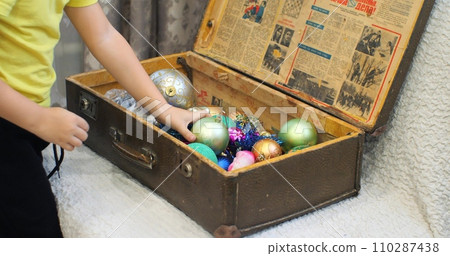 Children and mother put New Year's toys in an old suitcase. Close-up of hands putting New Year's toys into a box. Concept of the end of winter holidays and New Year, Christmas. 110287438