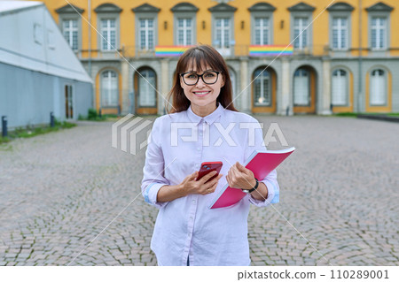 Middle-aged female teacher with smartphone, background of educational building 110289001