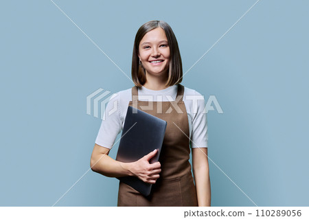 Portrait of young woman in apron holding laptop on blue background 110289056