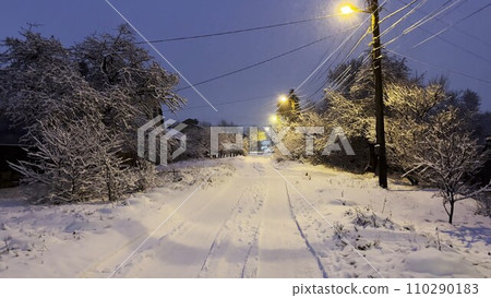 Snowy country road at evening time. Street lamp illuminating snow trail at winter night. Beautiful nature landscape with falling snowflakes at background. 110290183