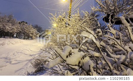 Close up of snow-covered tree branches illuminated by street lamp at evening time. View on country snowy road at winter night. Beautiful nature landscape at background 110290185