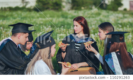 Graduates in black suits eating pizza in a city meadow. Graduates in black suits eating pizza in a city meadow. 110291263