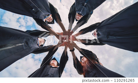 Team of college or university students celebrating graduation. Group of happy successful graduates in academic hats and robes standing in circle and putting their hands together. 110291265
