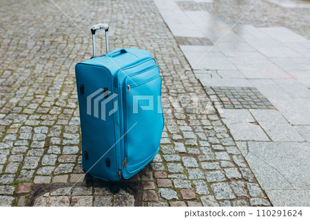 Travel luggage of empty European street. Tourism, Blue fabric suitcase on the cobbled street, rainy weather. Concept for travel or lost luggage 110291624