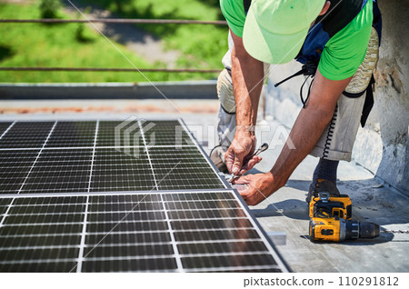 Worker building photovoltaic solar panel system on rooftop of house. Man engineer installing solar module with help of wrench outdoors. Alternative and renewable energy generation concept. Worker building photovoltaic solar panel system on rooftop of house. Man engineer installing solar module with help of wrench outdoors. Alternative and renewable energy generation concept. 110291812