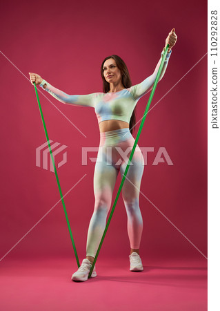 Confident female with dark hair, doing arm exercise with resistance band, while working out. Low angle view of fit woman in sportswear exercising, against pink background. Fitness and sport concept. 110292828