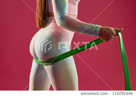 Close up of anonymous sexy female athlete exercising, strengthen buttocks with rubber band indoors. Crop view of seductive woman with green elastic band, working out, isolated on pink. Sport concept. 110292846