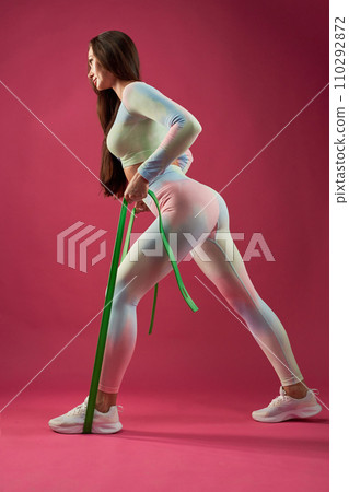 Determined sporty girl pulling green resistance rubber, while training in studio. Side view of determined stunning female athlete in gym outfit exercising, isolated on pink background. Sport concept. 110292872