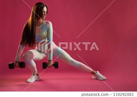 Strong brunette female in leggings stretching leg during fitness workout indoors. Front view of motivated middle-aged woman doing side lunge with dumbbells, against hot pink background. Sport concept. Strong brunette female in leggings stretching leg during fitness workout indoors. Front view of motivated middle-aged woman doing side lunge with dumbbells, against hot pink background. Sport concept. 110292881