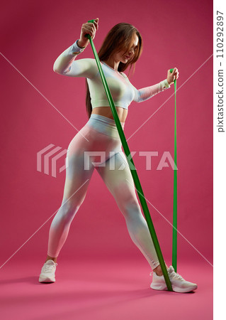 Slim Caucasian sportswoman stepping, pulling resistance rope, during fitness training indoors. Low angle view of focused young girl in sportswear, doing arm exercise, isolated on pink. Fitness concept 110292897