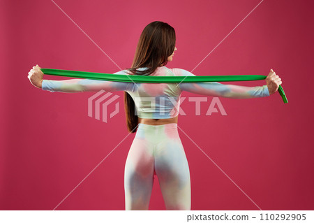 Attractive woman stretching rubber band behind back, while exercising indoors. Back view of long haired fit female with spread arms, doing rubber band exercises in studio. Fitness, lifestyle concept. 110292905