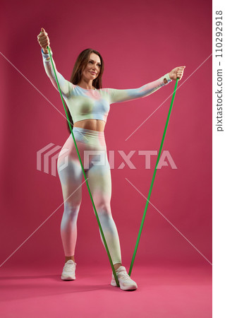 Female fitness trainer doing arm exercise with resistance band, working out indoors. Low angle view of attractive middle aged woman in legging exercising, against pink background. Fitness concept. Female fitness trainer doing arm exercise with resistance band, working out indoors. Low angle view of attractive middle aged woman in legging exercising, against pink background. Fitness concept. 110292918