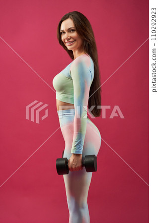 Beautiful fitness girl posing with dumbbell in studio. Side view of smiling caucasian sportswoman wearing activewear, looking at camera, while lifting dumbbell, against pink background. Sport concept. Beautiful fitness girl posing with dumbbell in studio. Side view of smiling caucasian sportswoman wearing activewear, looking at camera, while lifting dumbbell, against pink background. Sport concept. 110292923