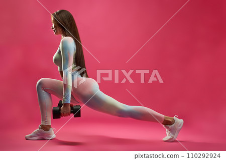 Athletic sexy woman in leggings stretching leg during fitness workout indoors. Side view of motivated sporty girl doing lunge with dumbbells, against hot pink background. Fitness, lifestyle concept. Athletic sexy woman in leggings stretching leg during fitness workout indoors. Side view of motivated sporty girl doing lunge with dumbbells, against hot pink background. Fitness, lifestyle concept. 110292924