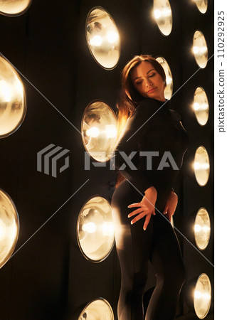 Gorgeous woman wearing black tight apparel, posing with hand on hips indoors. Side view of Caucasian beauty with closed eyes, demonstrating body shape in spot lights background. Female beauty concept. Gorgeous woman wearing black tight apparel, posing with hand on hips indoors. Side view of Caucasian beauty with closed eyes, demonstrating body shape in spot lights background. Female beauty concept. 110292925