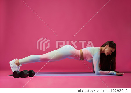 Focused brunette woman doing plank, working out in studio. Side view of shaped, fit young Caucasian girl exercising alone, isolated on bright pink background. Concept of healthy, sporty lifestyle. Focused brunette woman doing plank, working out in studio. Side view of shaped, fit young Caucasian girl exercising alone, isolated on bright pink background. Concept of healthy, sporty lifestyle. 110292941
