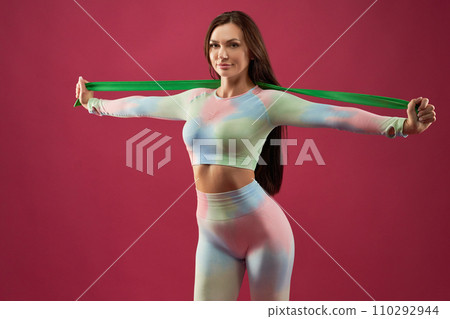 Strong, beautiful woman doing rubber band exercises in studio. Portrait of smiling female athlete with spread arms, exercising with elastic band, looking at camera, isolated on pink. Sport concept. 110292944