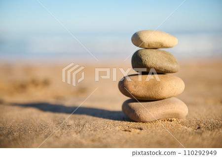 Still life with a pyramid of balanced stones stacked on the sand against sea background. Stacked pebbles. Copy ad space Still life with a pyramid of balanced stones stacked on the sand against sea background. Stacked pebbles. Copy ad space 110292949