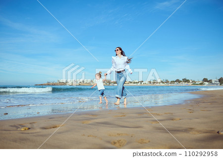 Mother and daughter holding hands, running barefoot on the wet sand, leaving footsteps, enjoying happy moments together 110292958