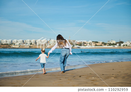 Little girl holding her mother's hand while walking in the warm water along the sea, enjoying the waves washing her feet 110292959