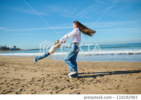 Full length portrait of a happy mother spinning her little child girl, an adorable daughter while playing on the beach 110292965