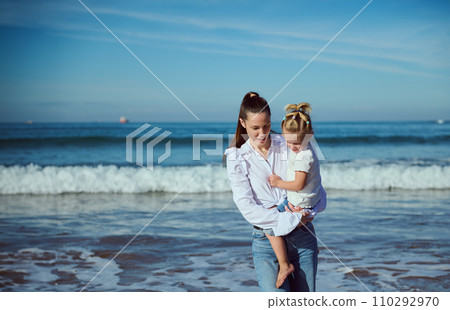 Authentic young woman talking with her daughter while carrying her, walking together on the beach 110292970