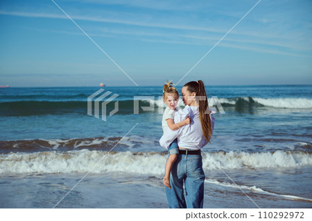 A beautiful young mother carries her baby in her arms, an adorable little girl, walking together barefoot on the beach. 110292972