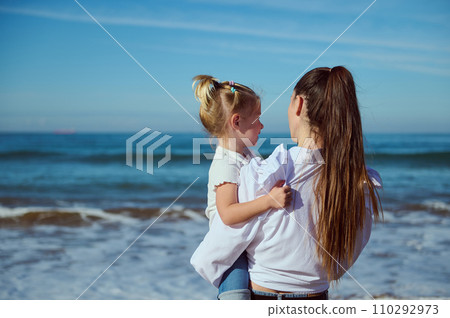 Rear view of mother holding her daughter and admiring waves splashing while pounding on the sandy shore. Family pastime 110292973