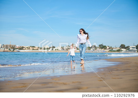 Mother and daughter holding hands and walking barefoot on the splashing waves of tropical sandy beach Mother and daughter holding hands and walking barefoot on the splashing waves of tropical sandy beach 110292976
