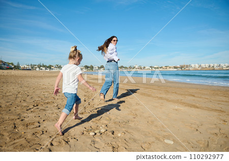 Rear view of young mother and her daughter, playing together on the beach, running and leaving footsteps on the wet sand 110292977