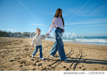Young mother and daughter holding hands and walking along the tropical beach, enjoying a happy weekend together outdoor Young mother and daughter holding hands and walking along the tropical beach, enjoying a happy weekend together outdoor 110292978