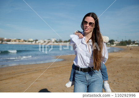 Happy Caucasian young mother carrying her little daughter on her back at the beach under a blue sky in the morning Happy Caucasian young mother carrying her little daughter on her back at the beach under a blue sky in the morning 110292996