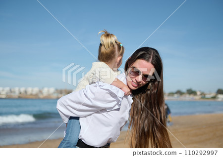 Authentic family of one young mom carrying her cute child girl on her back, enjoying a happy day on the beach Authentic family of one young mom carrying her cute child girl on her back, enjoying a happy day on the beach 110292997
