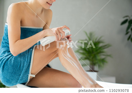 Slim anonymous female applying body lotion on one leg in bathroom. Crop view of smiling girl wrapped in turquoise cotton towel, using soap cream to wash in morning. Skin care, routine concept.  110293078